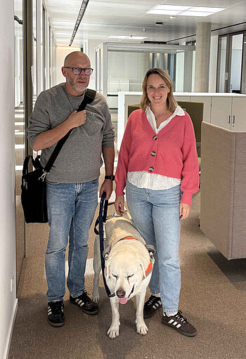 Ein Mann mit Blindenführhund und eine Frau in Büroräumen.